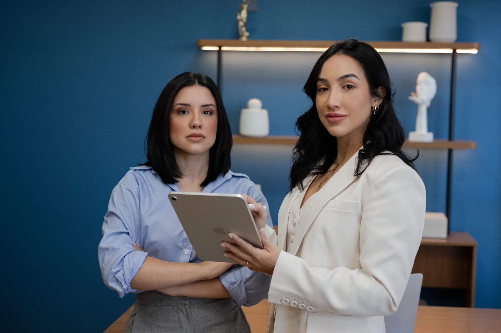 Vanessa Azevedo e Bárbara Feltrim no escritório, diante da estante na parede azul